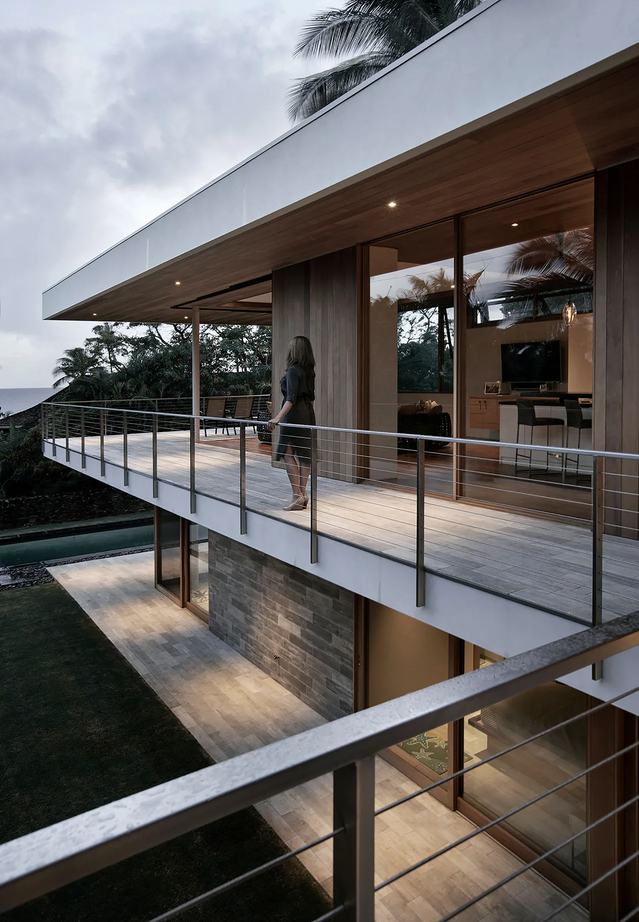 Woman standing on a second-floor balcony of a modern house with wood and stone exterior details at dusk.