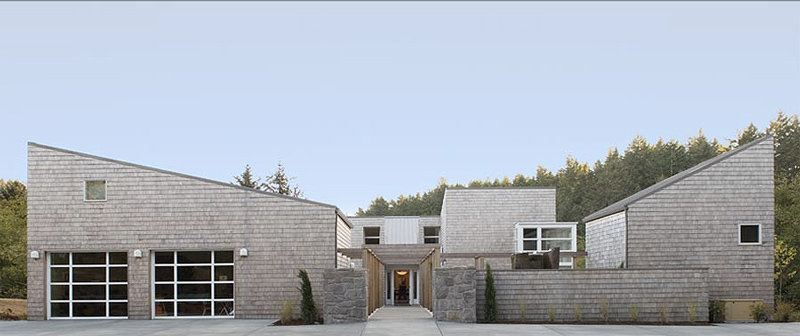 Front exterior view of a modern cabin with multiple gable roofs, shingle siding, stone walls, and a concrete driveway leading to the garage.