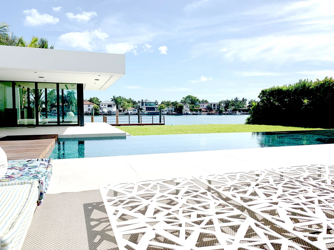 Modern house exterior with a flat roof, large glass windows, and an infinity pool overlooking a waterway and neighboring homes.