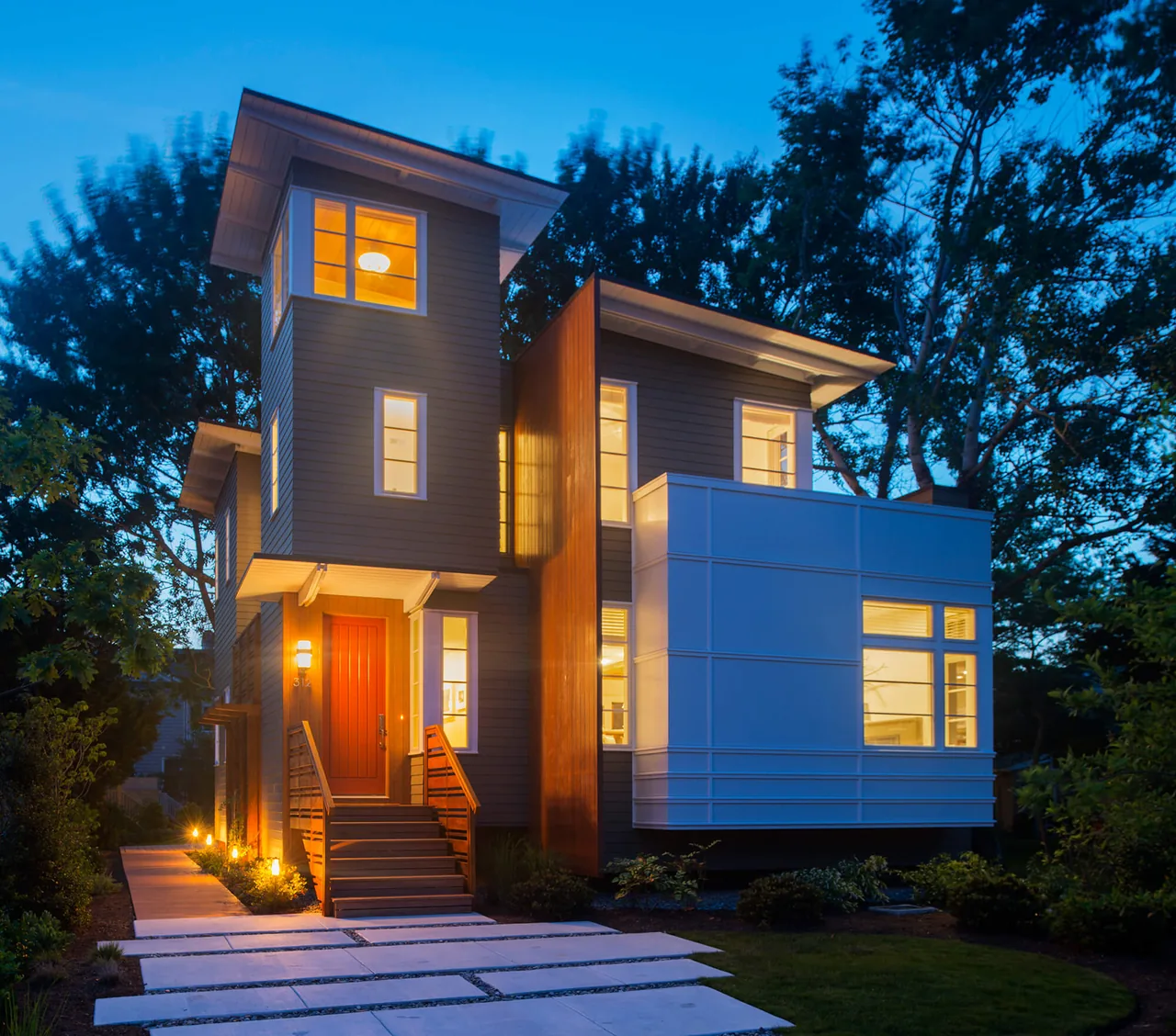 Evening view of a modern three-story home with warm interior lights and dark siding contrasted by a white panel section and a red front door