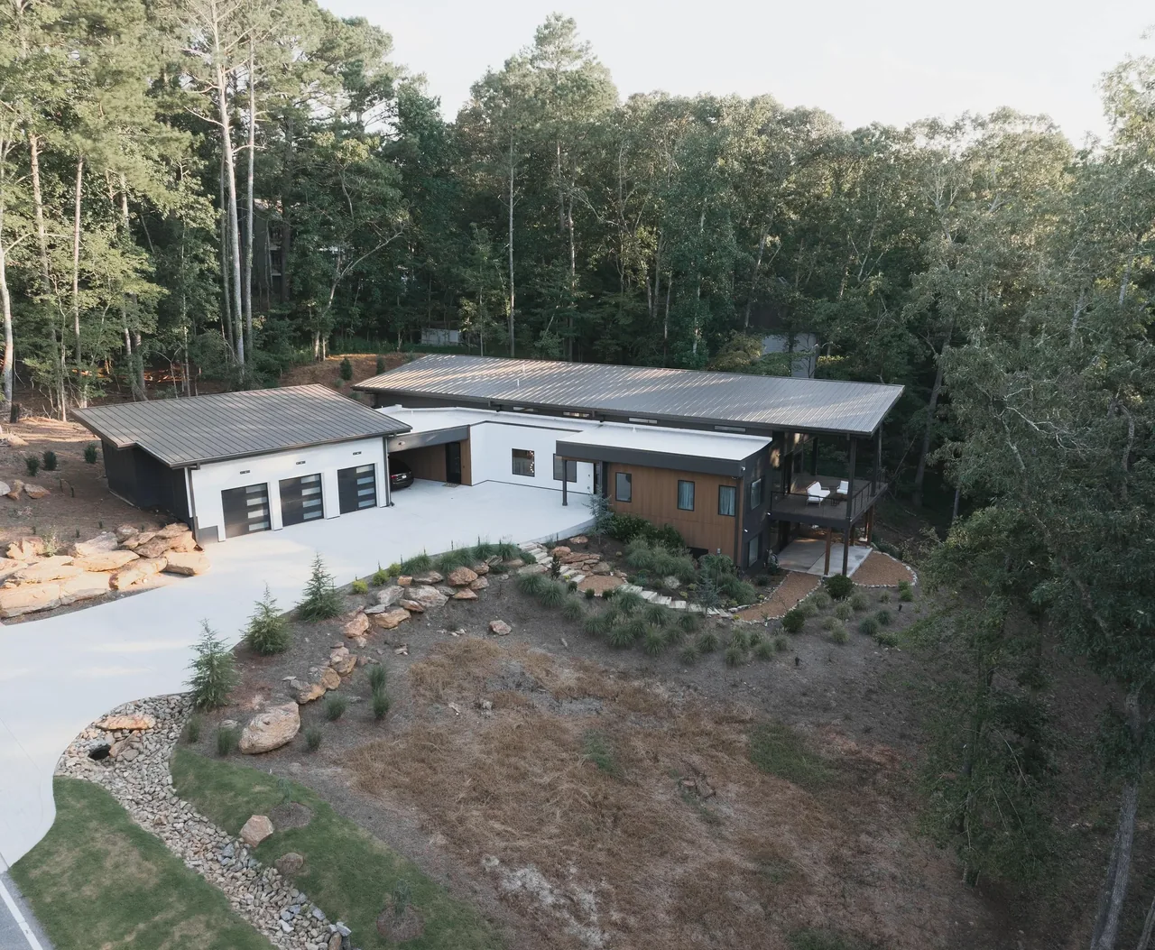 A modern flat-roof house with white, black, and wood siding situated in a wooded area with a rocky landscaped yard.