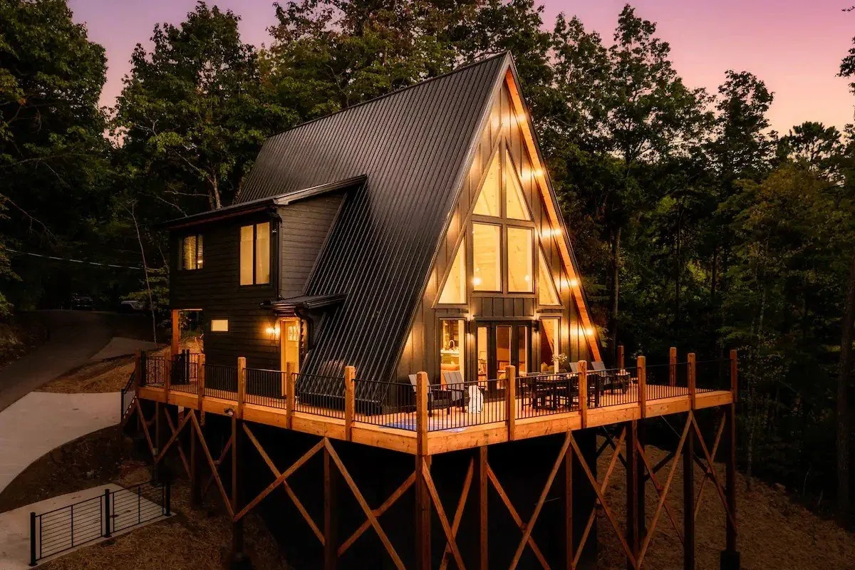A modern A-frame house elevated on wooden stilts with a large deck, glowing warmly at dusk surrounded by trees.