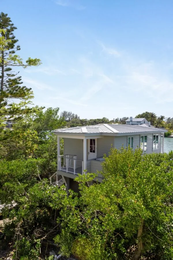 A single-story light gray house with a metal standing seam roof elevated above greenery and overlooking water.