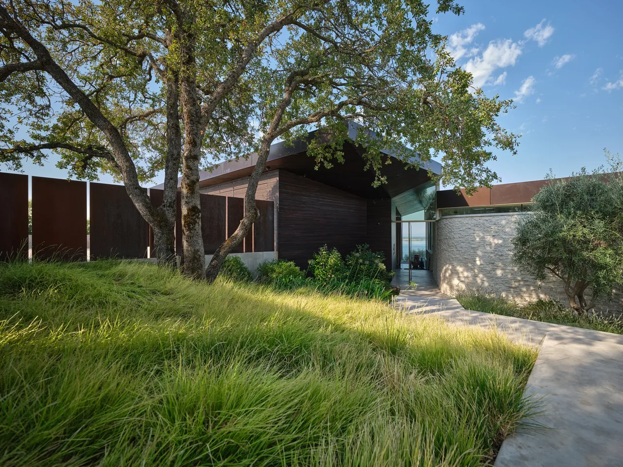 Modern house entry with wood siding, stone wall, and rust-colored steel panels surrounded by lush greenery and large trees.