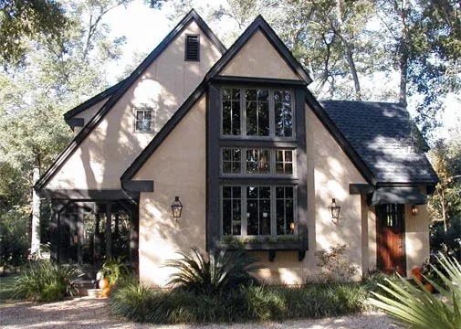 Front exterior view of a steep gable roof house with black window frames and stucco walls surrounded by trees.