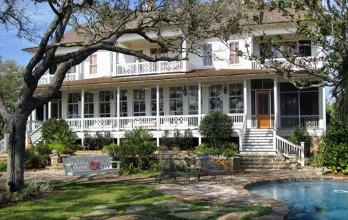 White two-story house with a large porch and a swimming pool in the foreground in a green yard with trees.