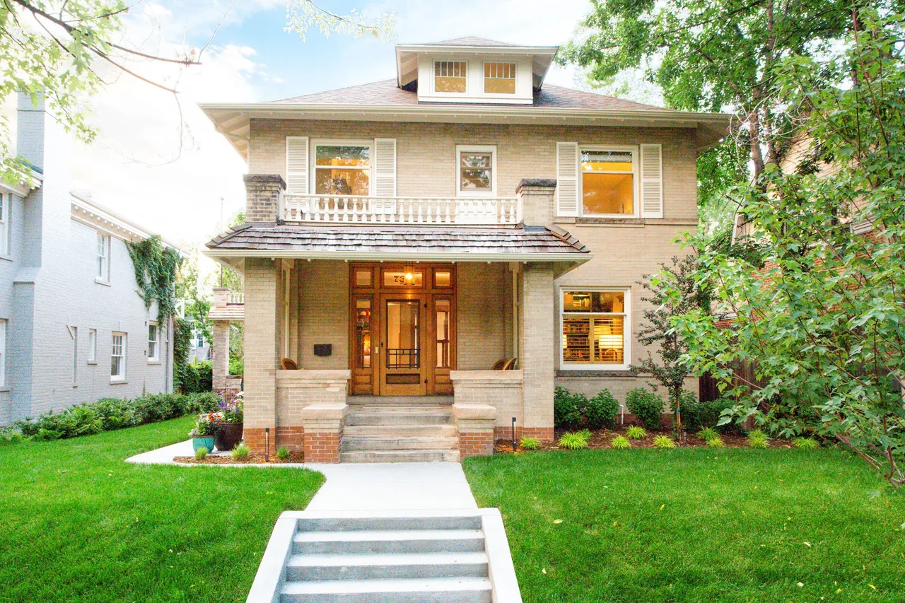 Front exterior view of a two-story beige brick house with a covered porch and wooden door framed by greenery.