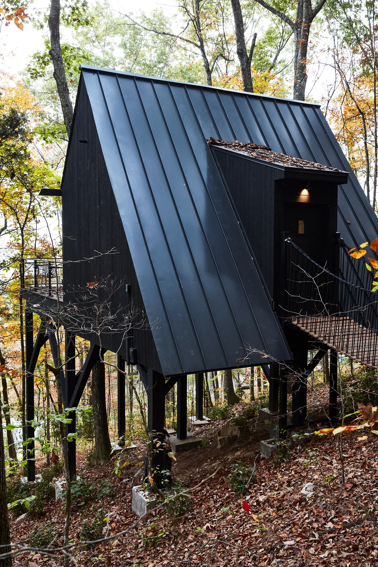 Elevated black A-frame house with a steep standing seam metal roof nestled in a wooded area in fall.