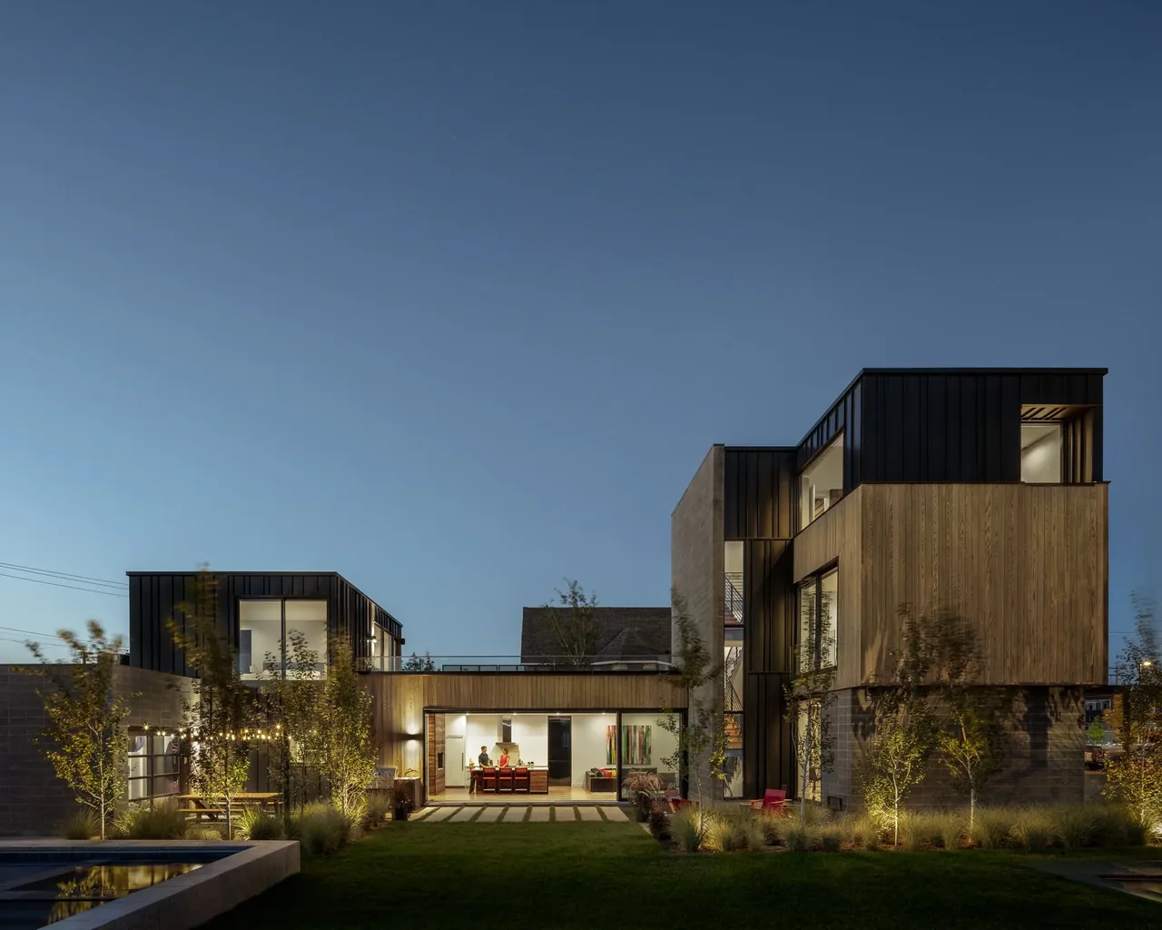 Evening view of a modern two-story residence with wood and black metal cladding, illuminated and showing large windows and an open patio.