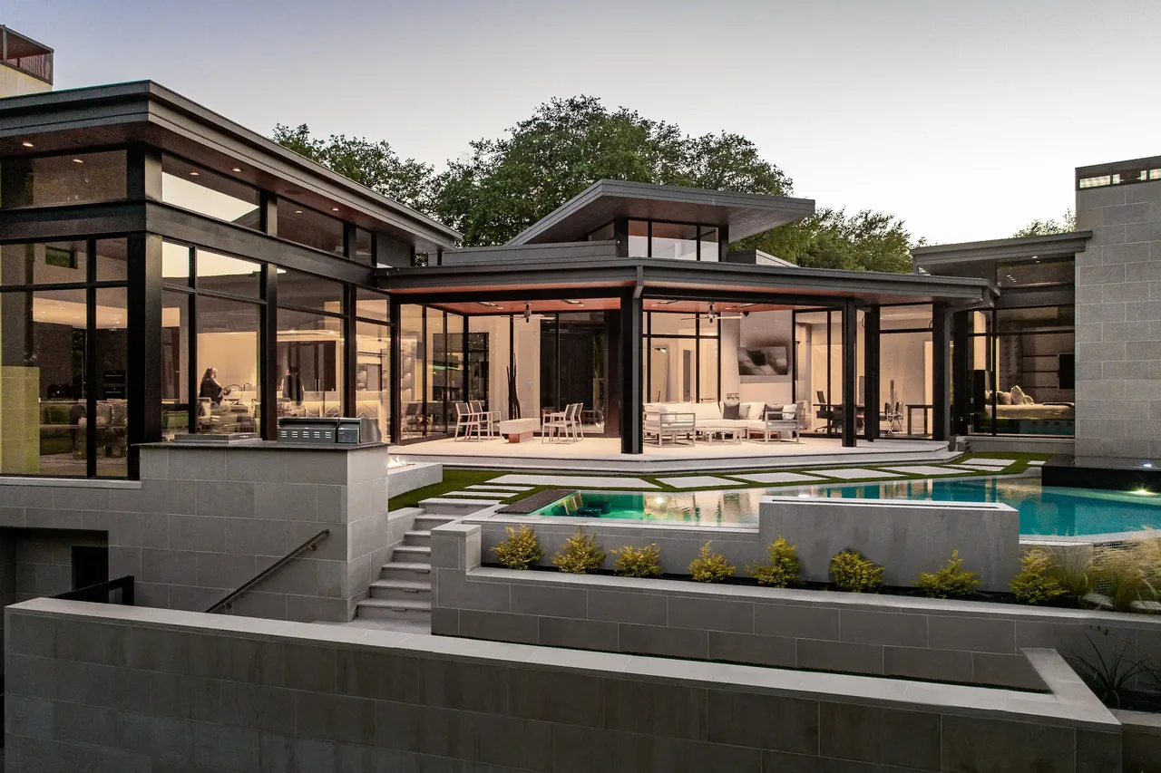 Exterior rear view of a modern home at dusk with large glass walls, covered patio, and swimming pool.