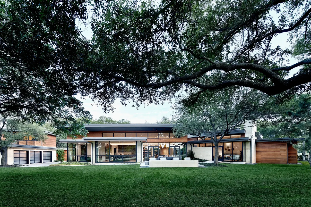 Rear exterior view of a modern one-story house with large windows and flat roof, surrounded by trees and green lawn.