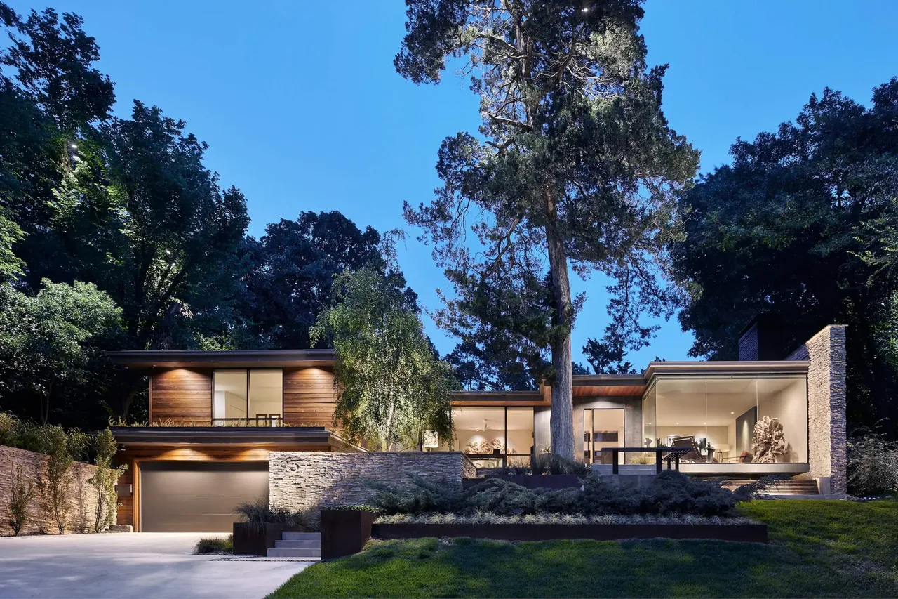 Twilight view of a modern two-story house with wood siding, stone walls, and large glass windows surrounded by trees.