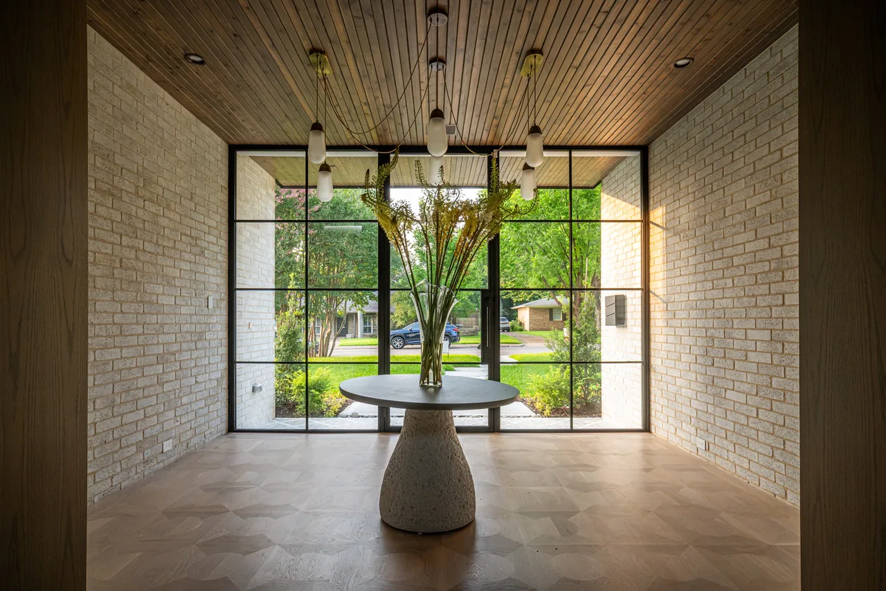 Entry space with a round table holding a tall vase of flowers in front of large black-framed grid windows looking out to a green yard.
