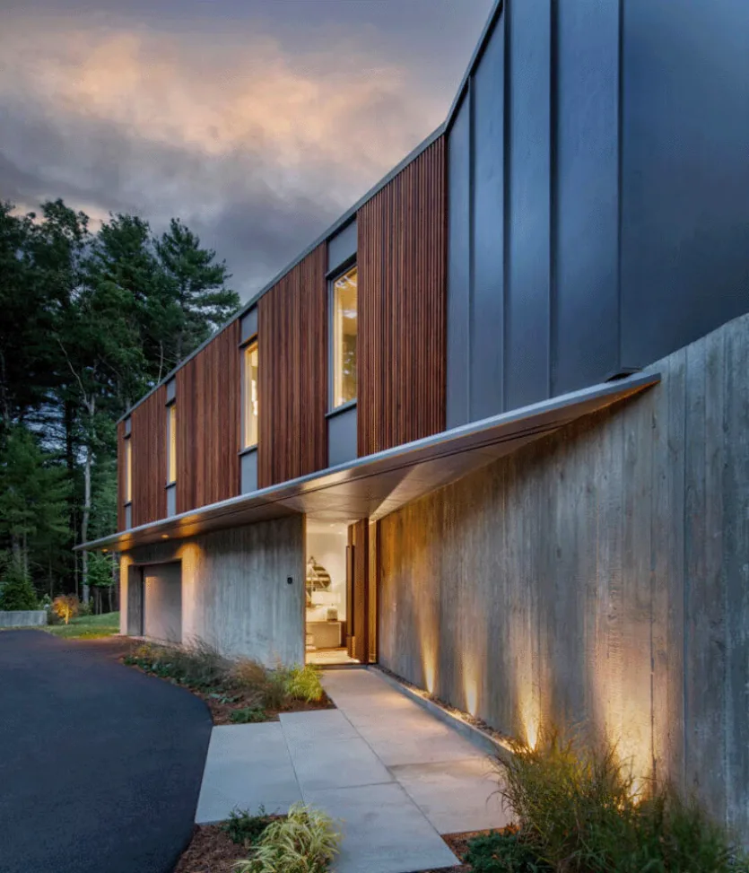 Exterior view of a modern home at dusk with a concrete lower wall and wooden vertical siding on the upper level beneath a gray metal roof.