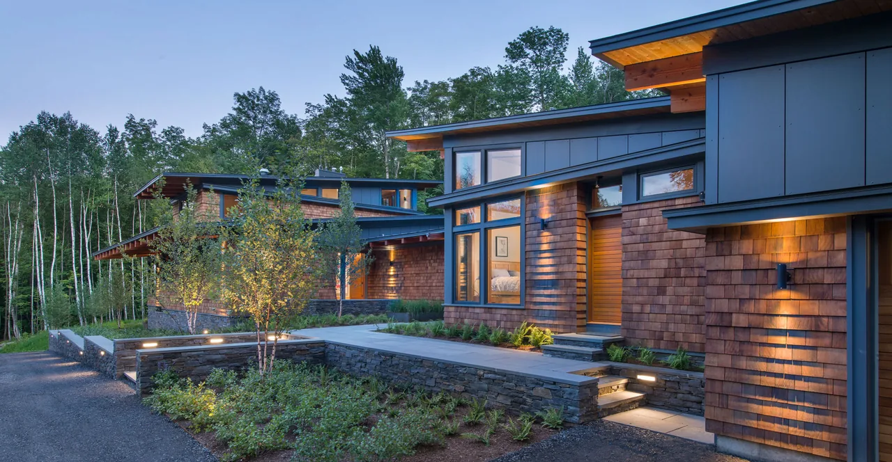 Modern house exterior with wood shingle siding, flat and shed roofs at dusk with exterior lighting.