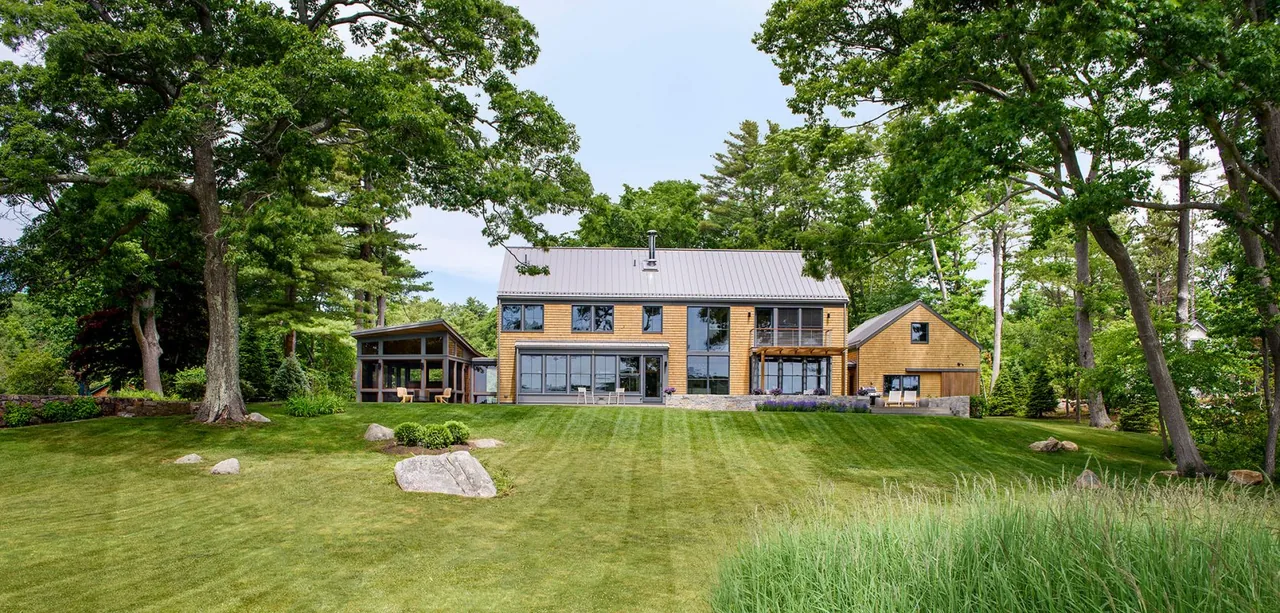Rear view of a modern farmhouse with wood siding and large windows, set in a green, wooded landscape.