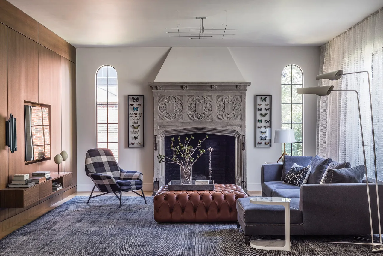 A living room with a gray sectional sofa, patterned armchair, and a detailed stone fireplace centered between two arched windows with sheer,