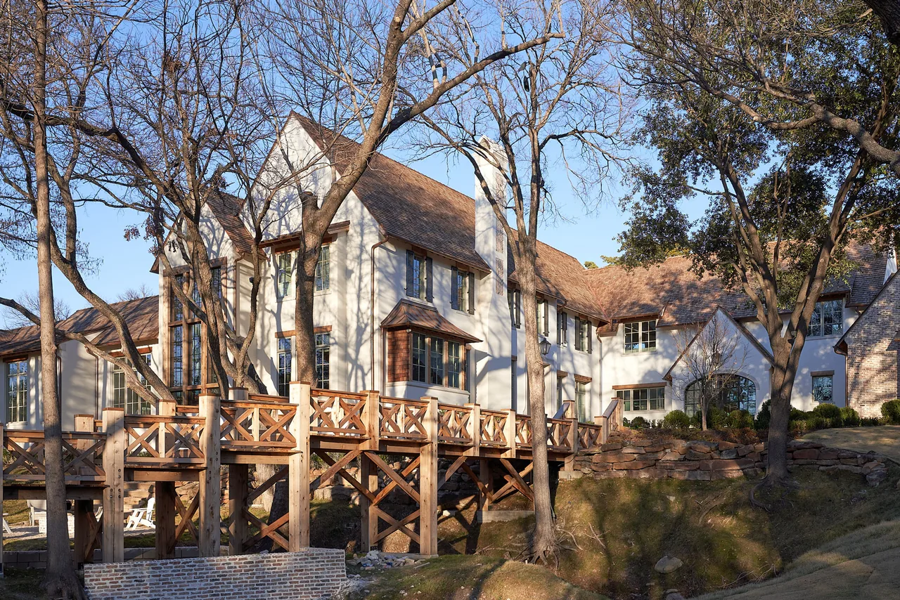 Exterior view of a large two-story house with white stucco walls, a steeply pitched roof, and a wooden elevated walkway surrounded by leaf-l