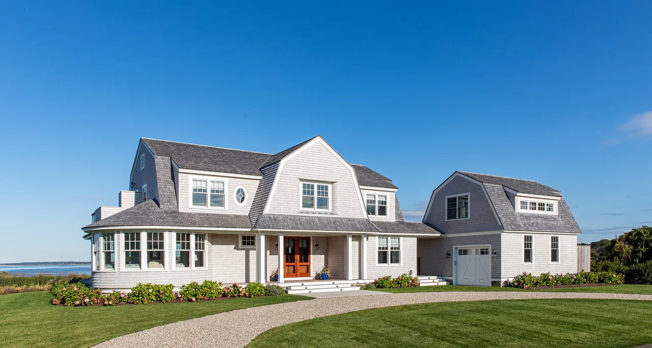 White shingle-sided house with detached garage under blue sky near a body of water.