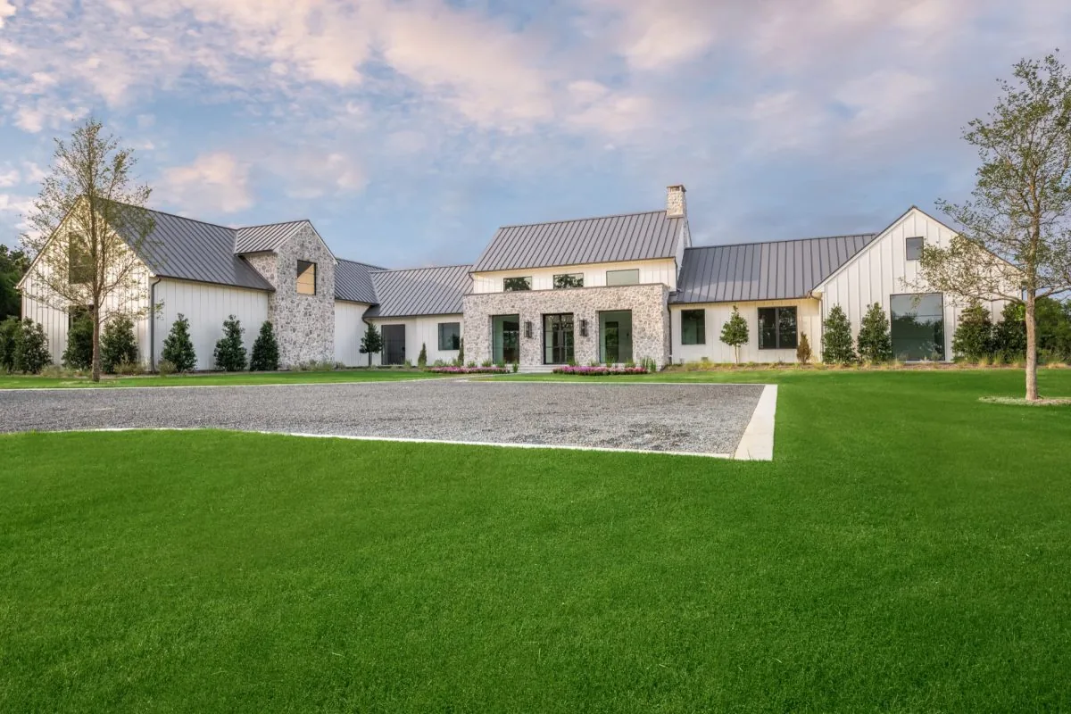 Front exterior view of a Hill Country Estate home with white siding, stone accents, and a metal roof under a partly cloudy sky.