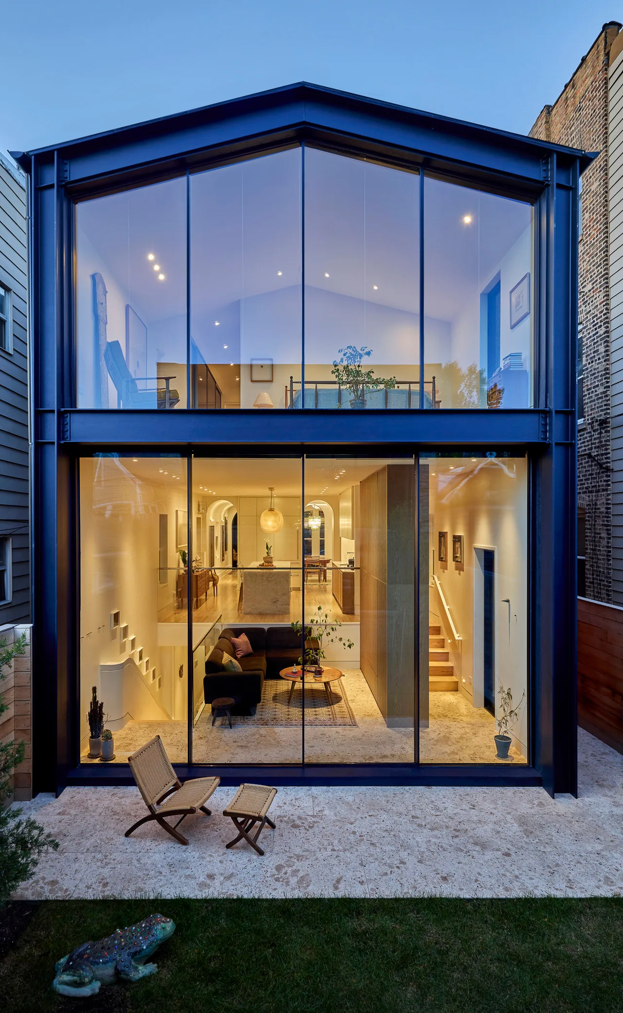 Rear exterior view of a modern two-story residence with large floor-to-ceiling glass windows revealing the interior living and dining areas.