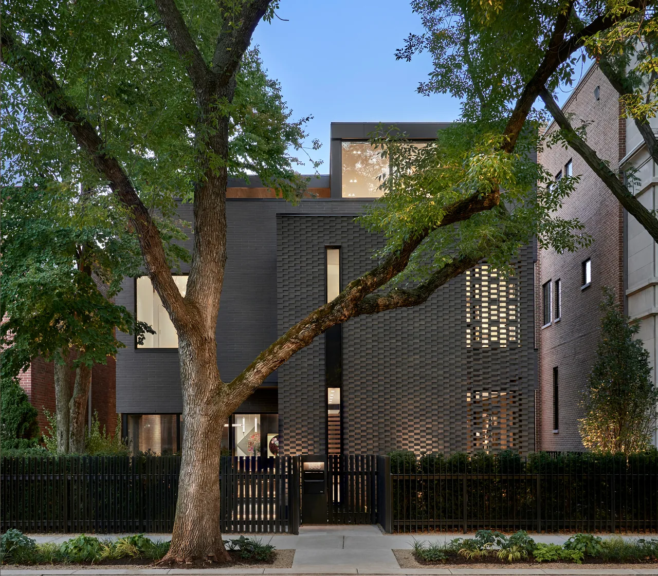 Dark brick modern house facade with a patterned brick wall and large tree in front at dusk.