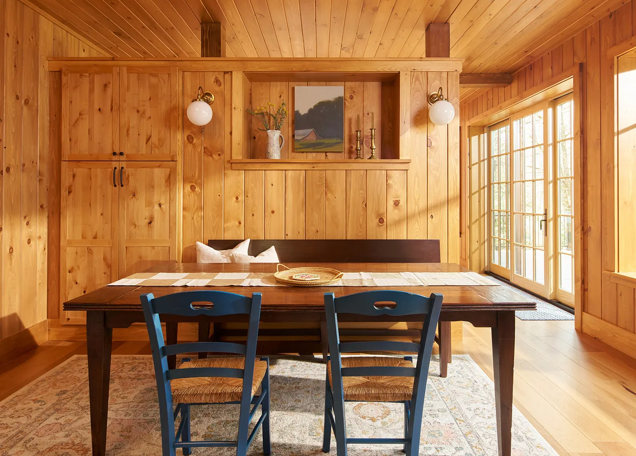 Dining area with wooden walls, ceiling, and floor, featuring a dark wooden table, blue chairs, and French doors letting in natural light.