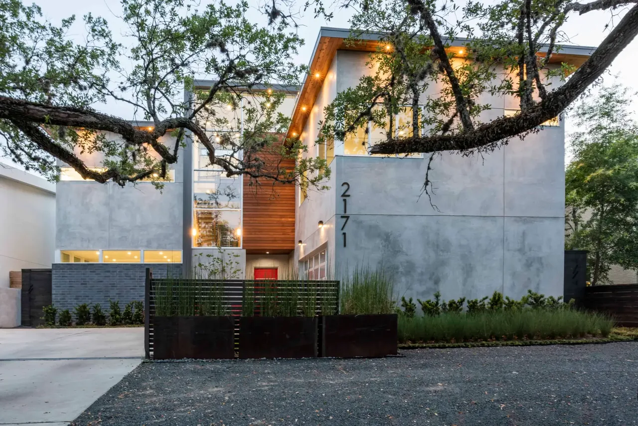 View of a modern two-story house exterior with concrete walls, black window frames, and desert landscaping at dusk.