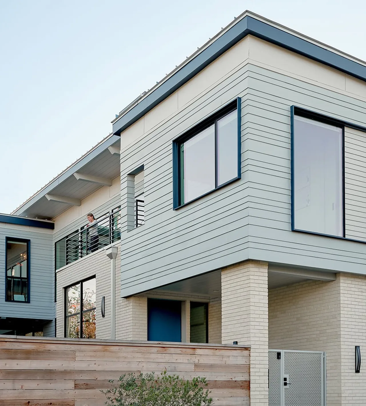 White and light gray modern two-story house with blue window frames and a wooden privacy fence.