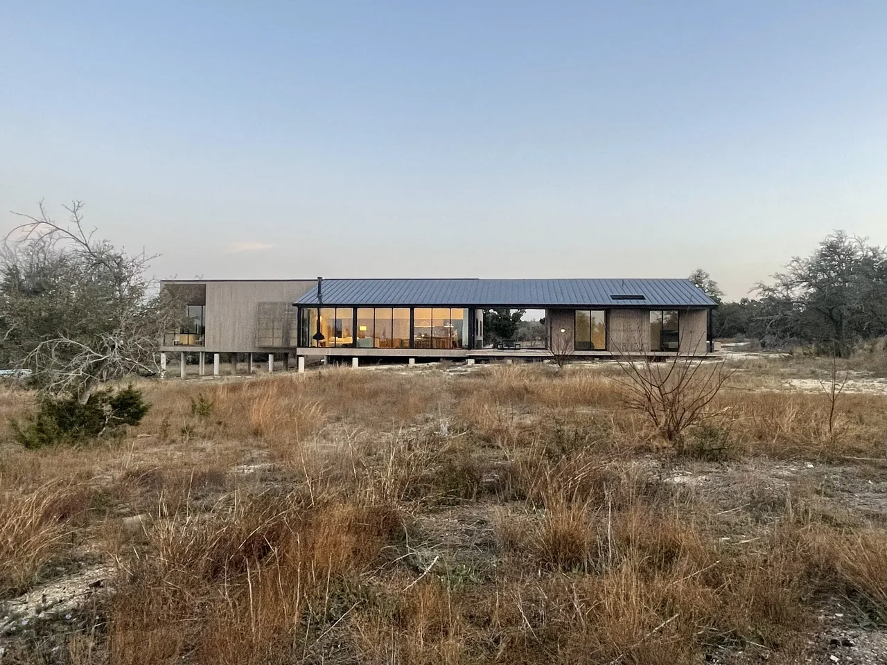 Long, elevated modern house with vertical wood siding, large windows, and a standing seam metal roof in a dry grassland setting.