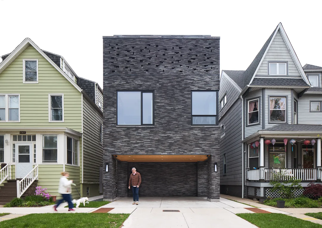 Front view of a modern box-shaped dark brick house flanked by traditional gable-roof houses on both sides.