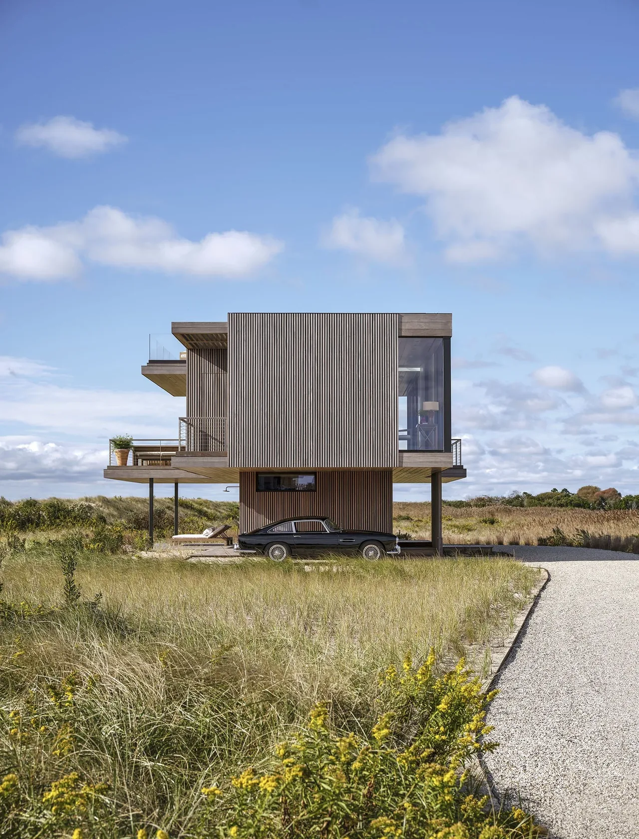 Modern two-story house on stilts with vertical wood siding surrounded by grass and a gravel driveway.