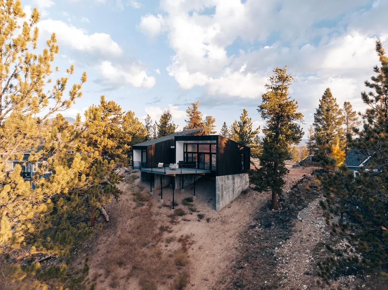 A modern house with dark wood siding and concrete foundation sits on a hillside surrounded by pine trees under a partly cloudy sky.