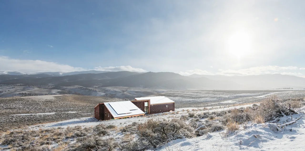 A modern house with redwood siding and skylights in a snowy, open valley under a bright sun.
