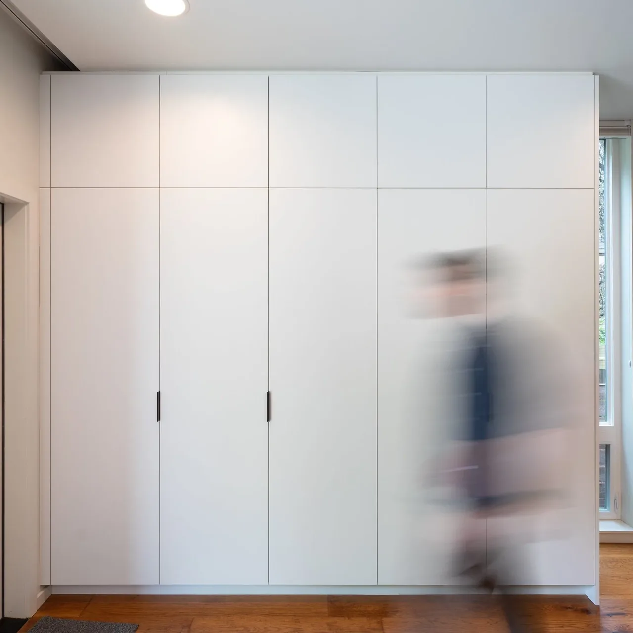 A tall white built-in cabinet with vertical handles in a room with wood flooring and a blurred person walking past.