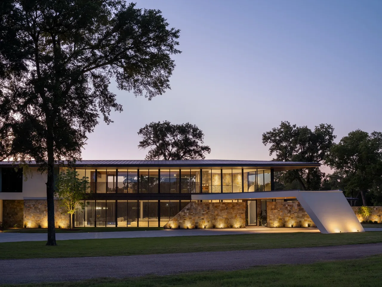 Modern two-story house with stone lower walls and extensive glass upper walls during dusk.