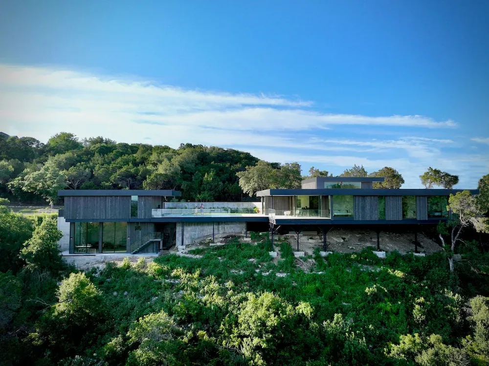 Modern house with multiple roof peaks perched on a hillside surrounded by trees under a blue sky.