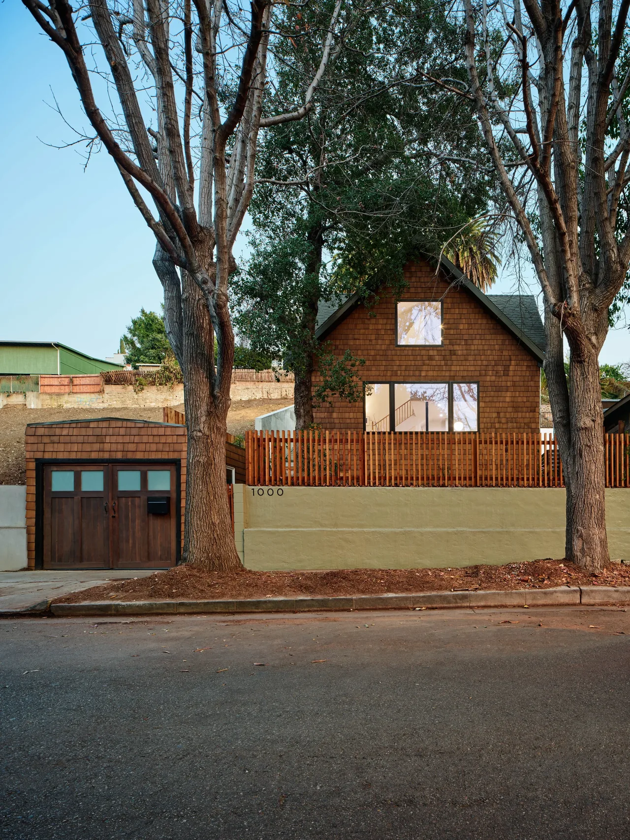 Exterior view of a small house with brown shingle siding and a detached garage behind a beige wall and wooden fence, flanked by two large,