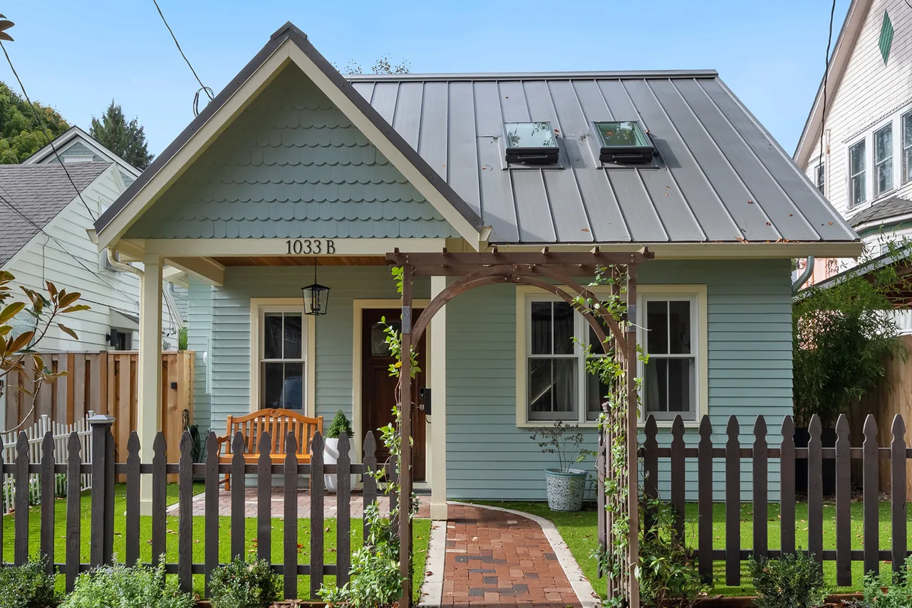 Small light blue house with a metal gable roof and a wooden picket fence with an arched entrance.