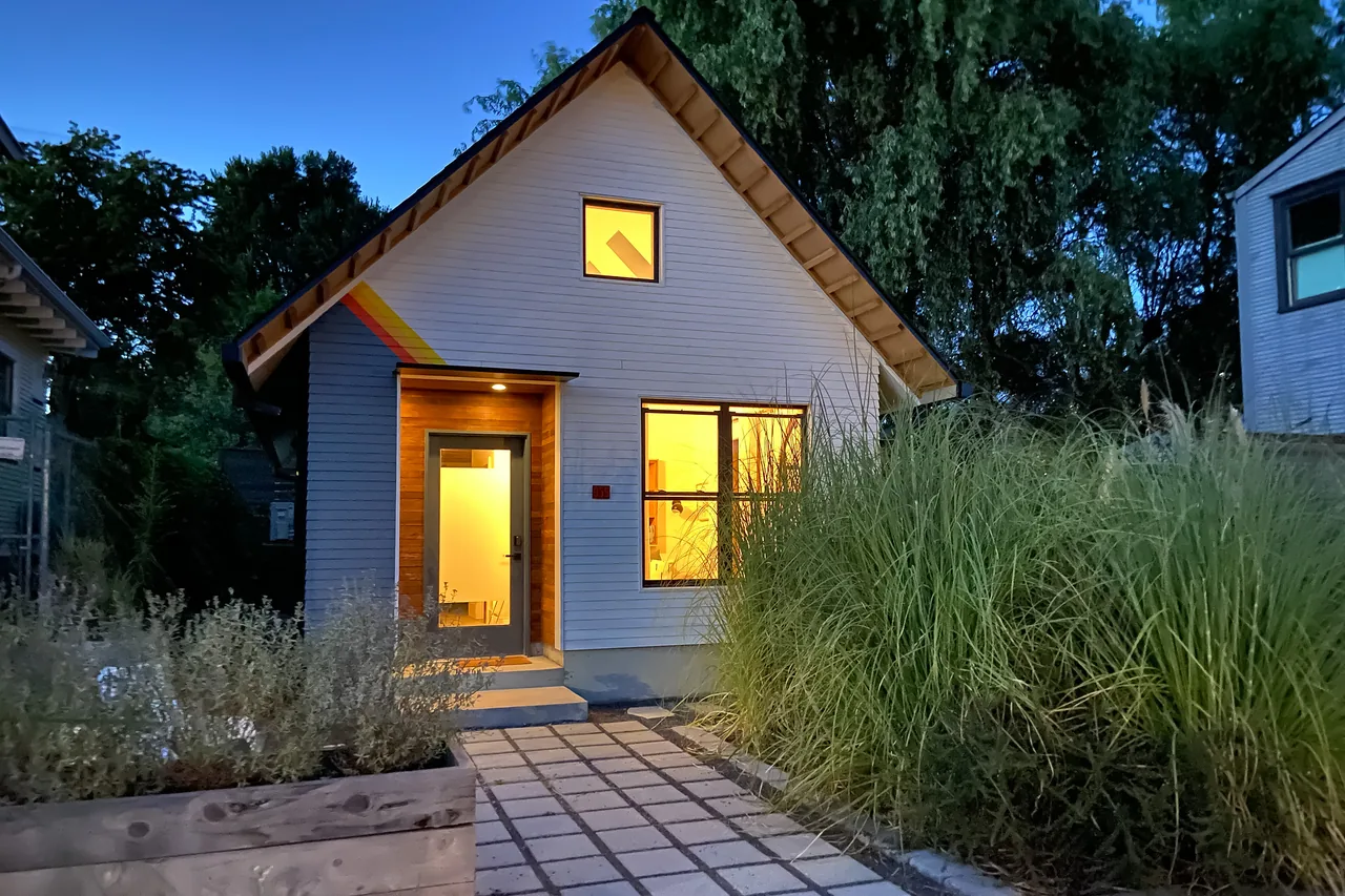 Small detached accessory dwelling unit at dusk with illuminated interior and white shingle siding, a steep gable roof, and colorful stripes.