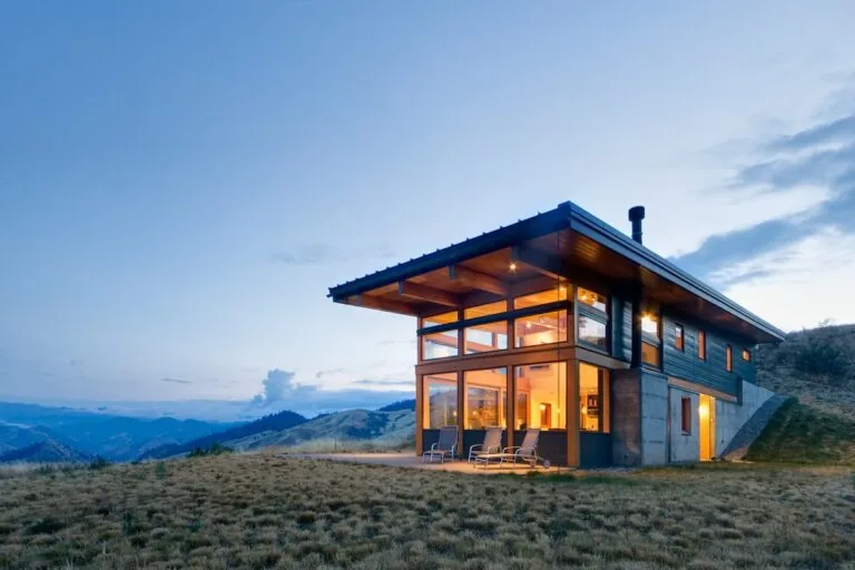 Modern house with large windows and wooden roof overlooking a grassy hillside at dusk.