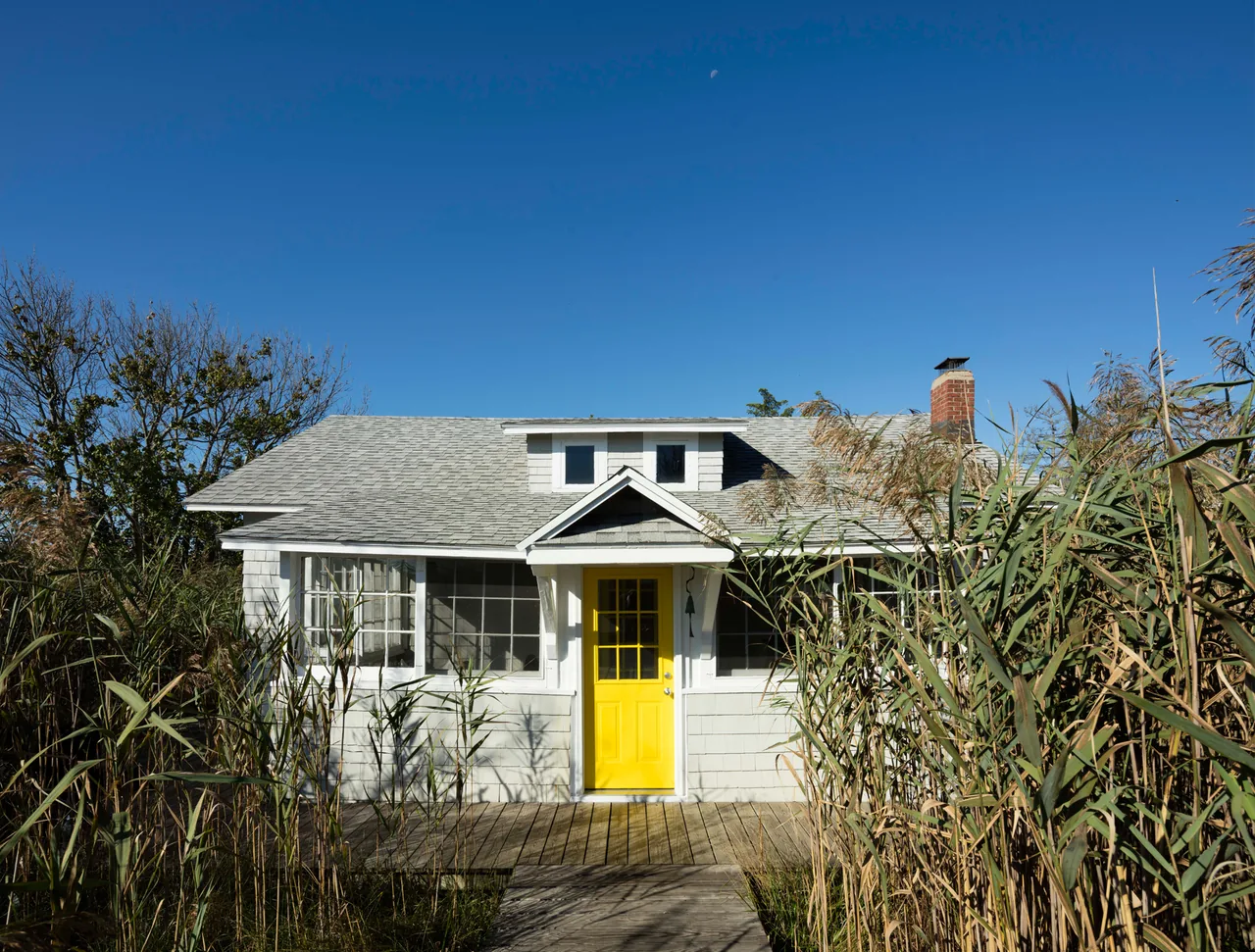 Small white house with a bright yellow front door surrounded by tall grasses under a clear blue sky.