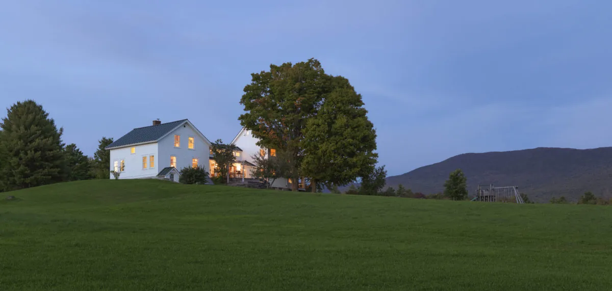 Distant view of a white two-story house with lights on, situated on a grassy hill with trees and mountains in the background at dusk.