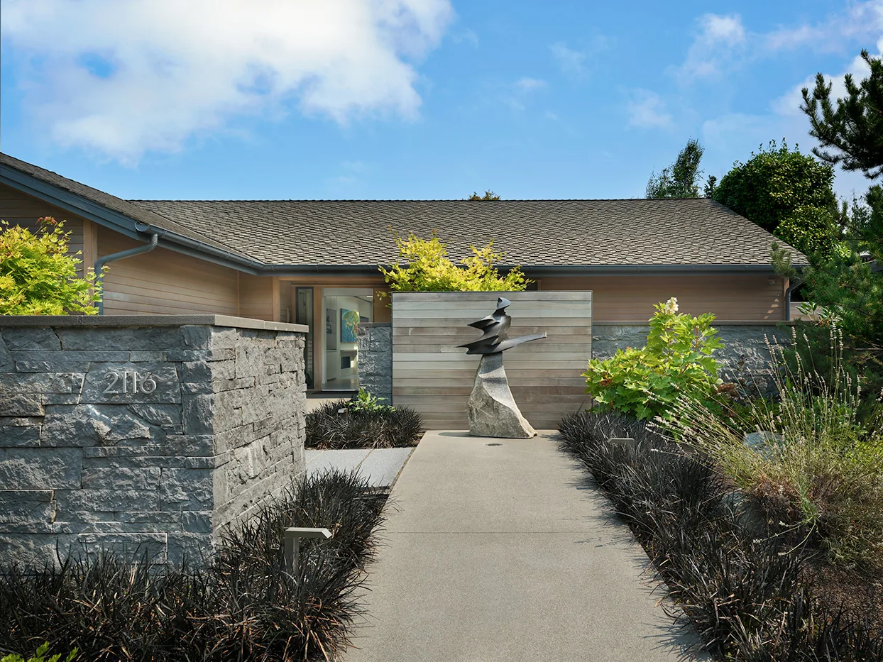 Front entrance pathway with stone walls and modern sculpture leading to a house with a shingled roof.