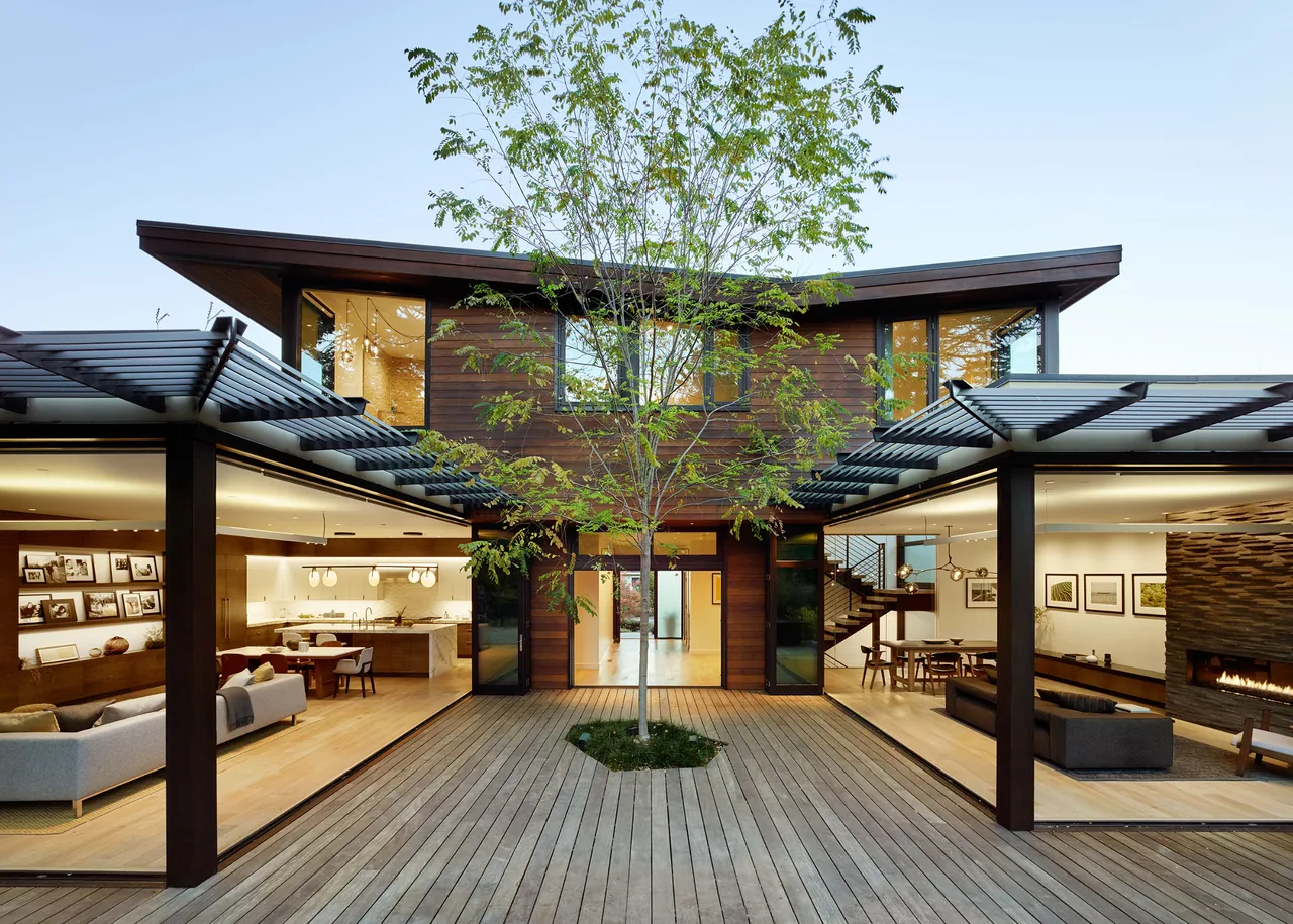 View of a modern two-story home with an open courtyard featuring a central tree and large folding glass doors.