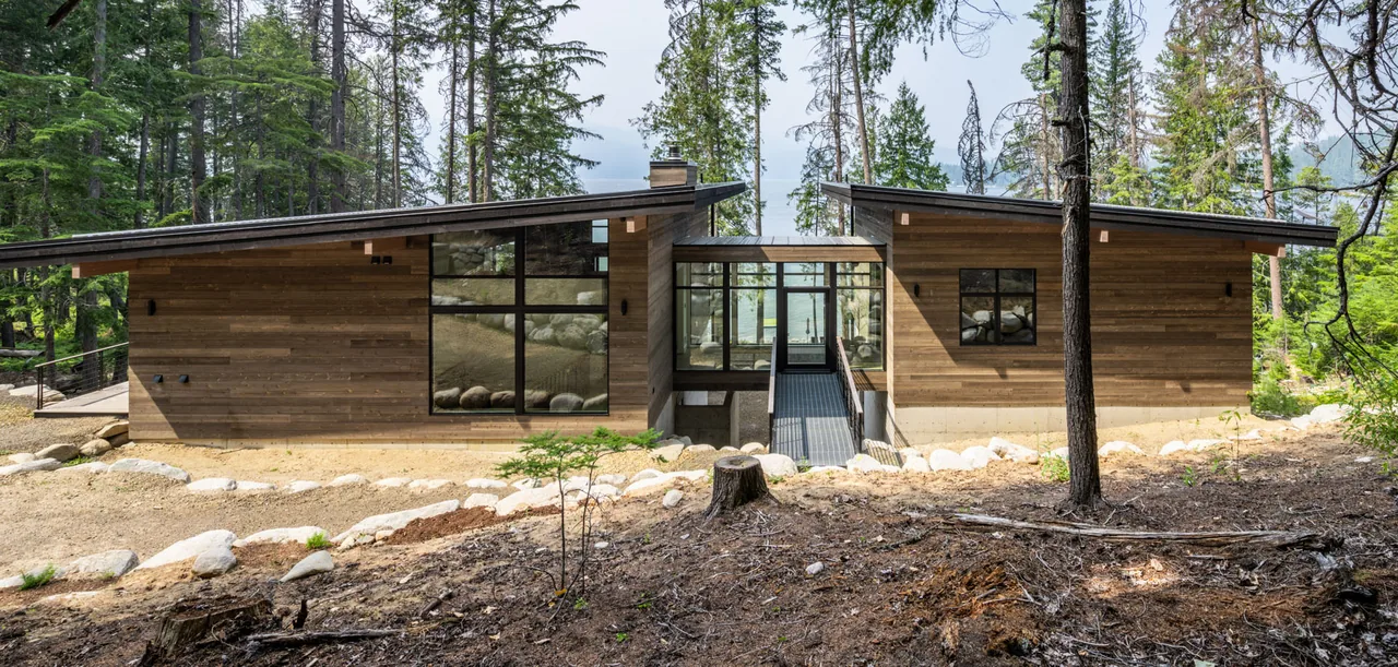 Single-story cabin with two sloped roofs connected by a glass hallway in a forest setting.