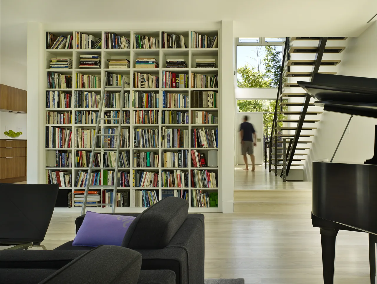 View of a modern living room with a large white built-in bookshelf filled with books, a dark gray sofa, and a black grand piano near a wide,