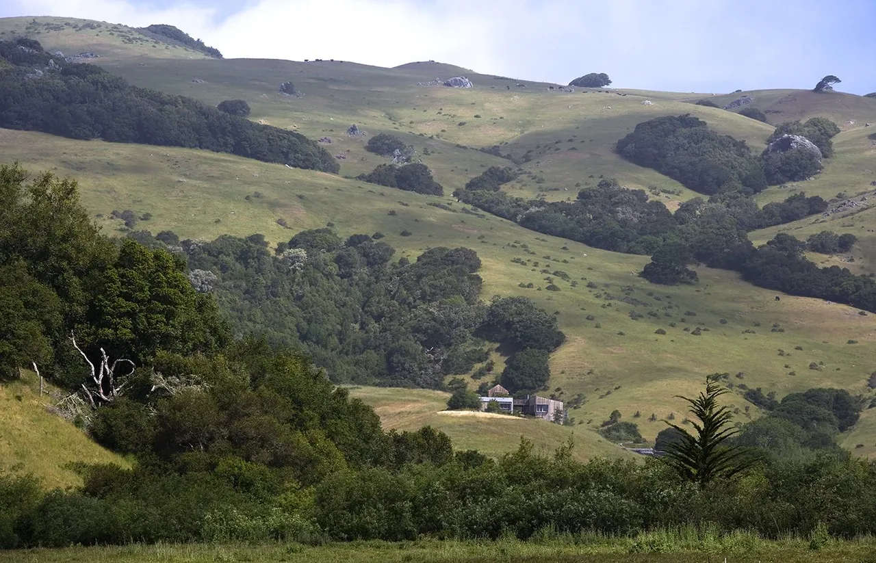 A ranch house sits nestled in green rolling hills with trees and rocky outcrops under a partly cloudy sky.