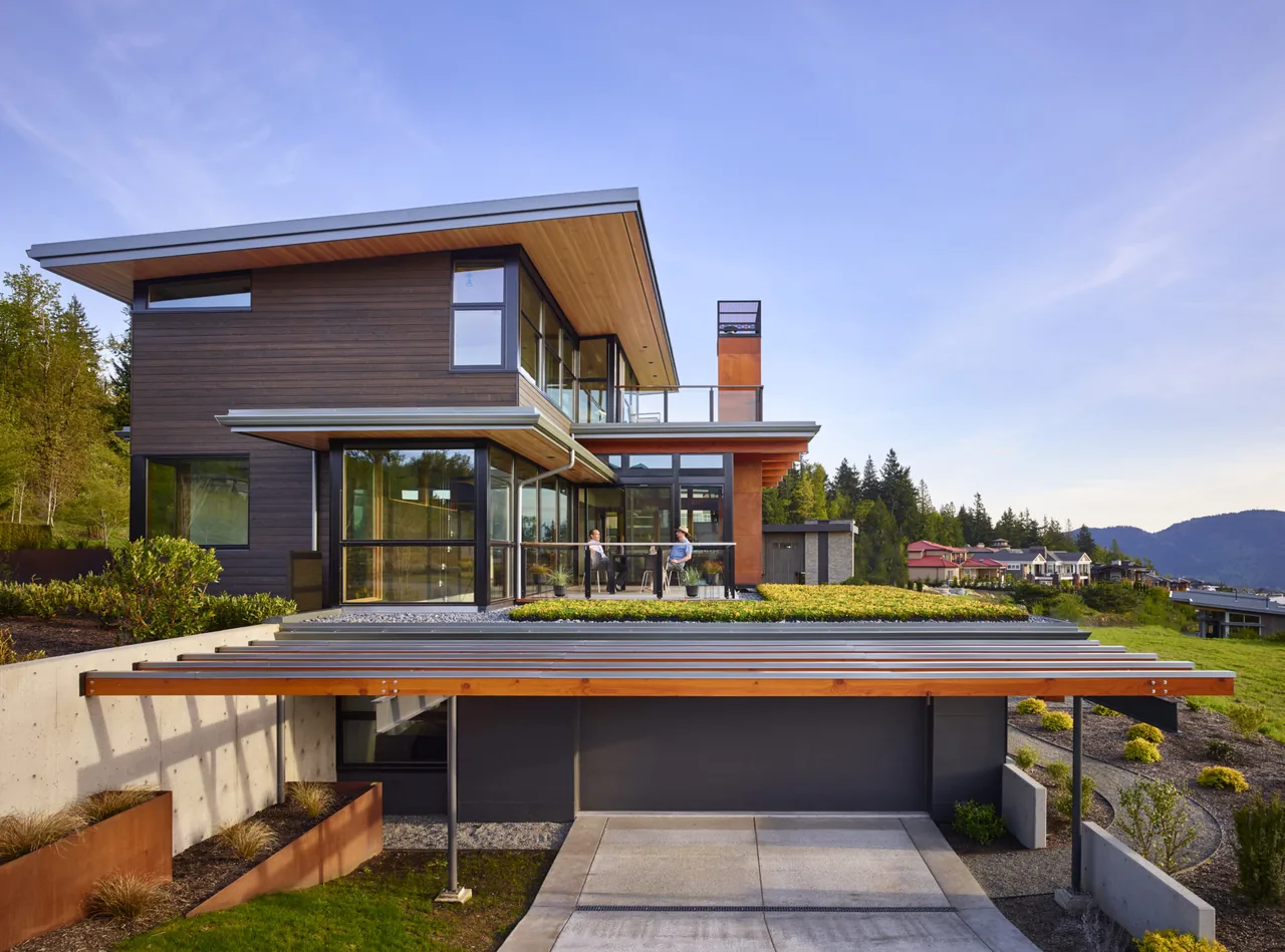 Modern two-story house with wood siding, large windows, and an angled roof, featuring a garage and green roof in a suburban setting.