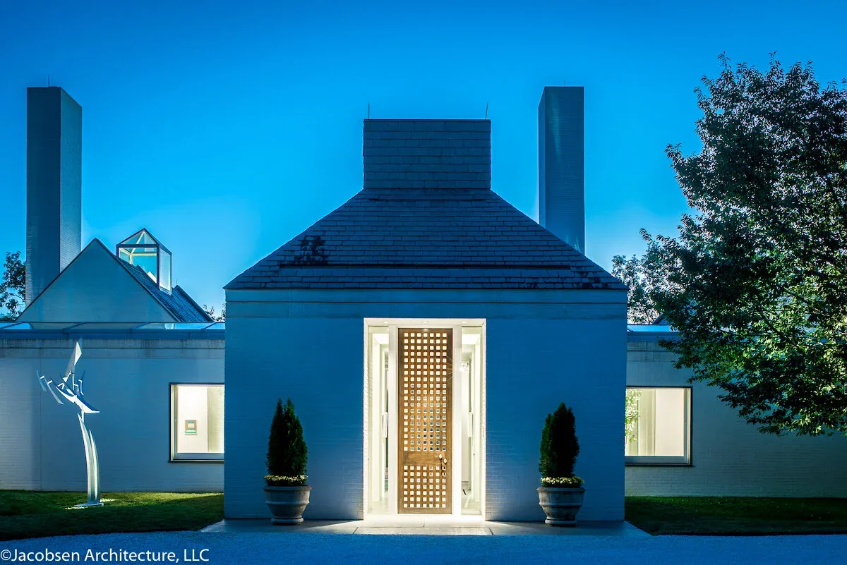 Evening view of a white brick modern house facade with a tall, unique wooden lattice front door and two potted plants on either side.